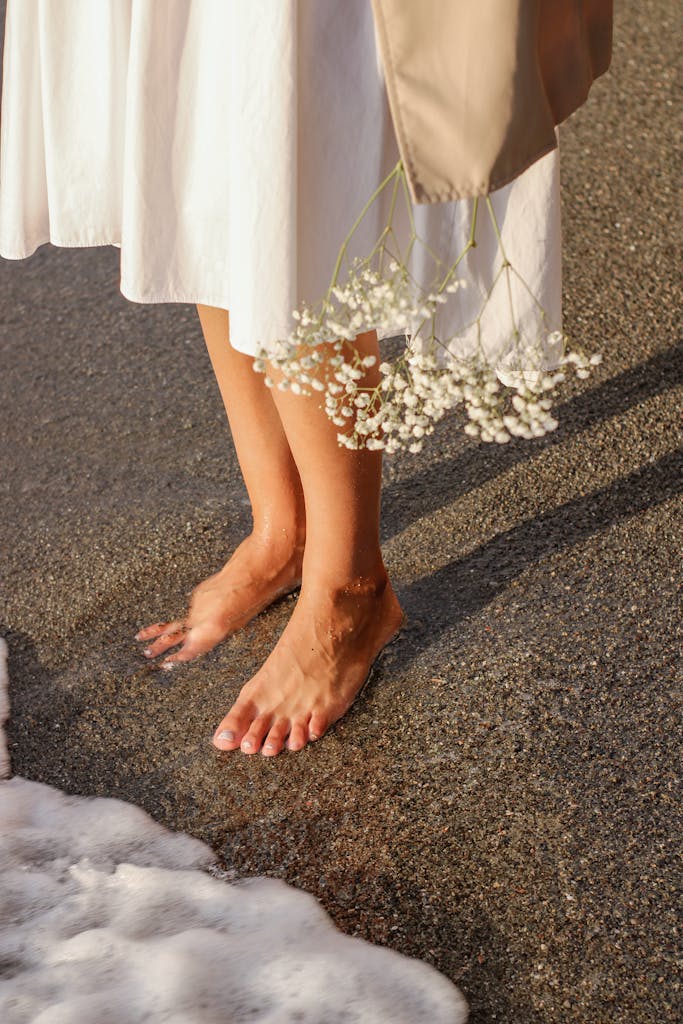 A woman in a white dress stands barefoot on the sandy shore holding flowers, capturing a serene beach moment.
